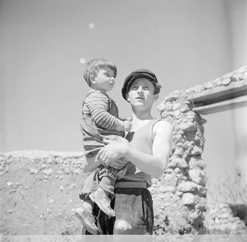 A young man holding a child by a stone wall, black and white scan